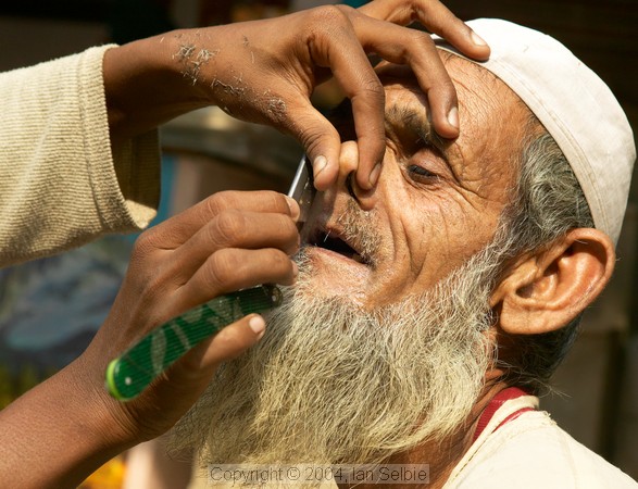 Young boy shaving the moustache of an old bearded man on the footpath by the street, Old Delhi