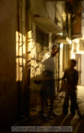 Nizammudin district, Delhi.  A side alley poorly lit, but with constant human traffic in the rather mysterious light cast by the windows of the adjacent house.
